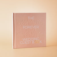 A hardcover wedding guest book with a matte blush pink cover, standing upright on a warm beige background. The cover features embossed white text reading 'THE FOREVER WEDDING GUEST BOOK' with 'PERO, MERON, MERON!' highlighted in white and gold.