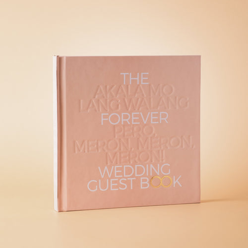 A hardcover wedding guest book with a matte blush pink cover, standing upright on a warm beige background. The cover features embossed white text reading 'THE FOREVER WEDDING GUEST BOOK' with 'PERO, MERON, MERON!' highlighted in white and gold.