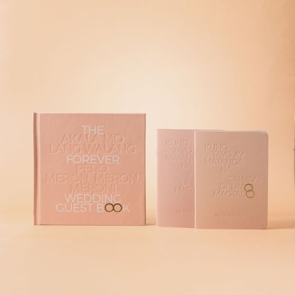 A blush pink wedding guest book set on a warm beige background, featuring a larger book with stacked text on the front and a smaller matching notebook beside it.